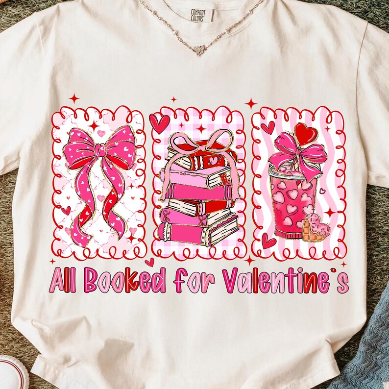 May include: Cream-colored t-shirt with a Valentine's Day graphic. The graphic features a pink bow, a stack of books, and a pink drink, all within decorative frames. The text "All Booked for Valentine's" is printed below.