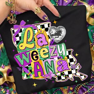 May include: Black t-shirt with a colorful graphic that says "La Weezu Ana". The design includes a disco ball heart, a crown, and a checkered pattern. The letters are in yellow, green, and purple.