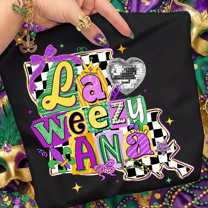 May include: Black t-shirt with a colorful graphic that says "La Weezu Ana". The design includes a disco ball heart, a crown, and a checkered pattern. The letters are in yellow, green, and purple.