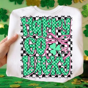 May include: White sweatshirt with the text "Happy Go Lucky" in green checkered letters with pink shamrocks. A pink bow and shamrock graphic are also featured. The design is set against a black and white checkered background.