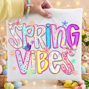 May include: White pillowcase with the words "SPRING VIBES" in colorful, whimsical lettering. The design includes flowers, bows, and a butterfly. The pillowcase is held up by a person wearing a bracelet.