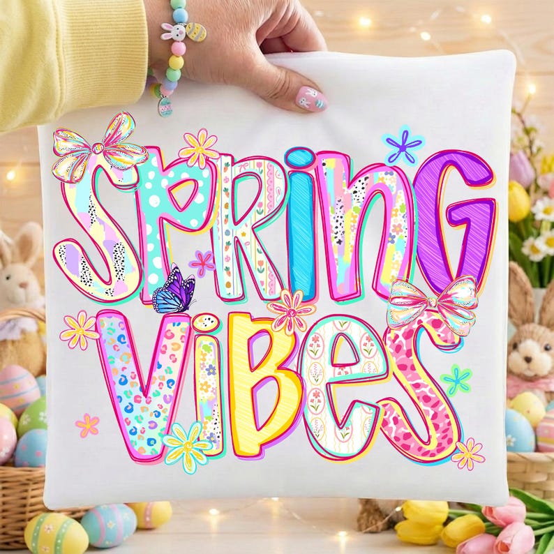 May include: White pillowcase with the words "SPRING VIBES" in colorful, whimsical lettering. The design includes flowers, bows, and a butterfly. The pillowcase is held up by a person wearing a bracelet.