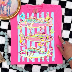 May include: A pink t-shirt featuring a colorful illustration of three fish. The fish are in shades of yellow, pink, and blue, set against light blue vertical stripes. Floral accents and the text "Comfort Colors" are visible.