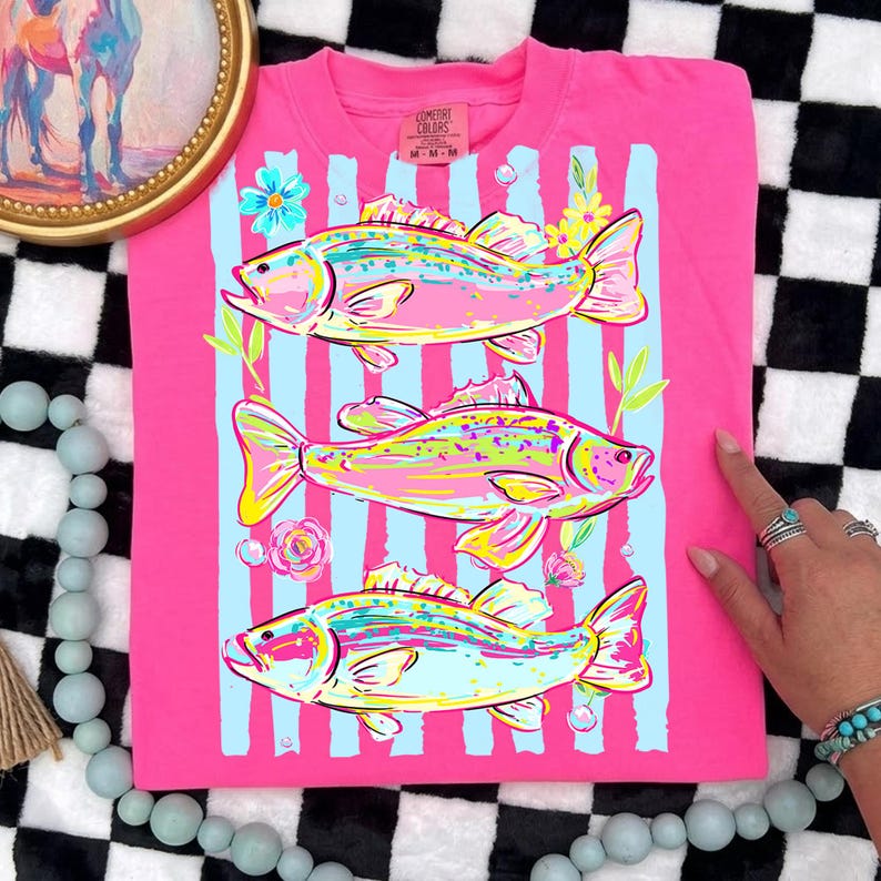 May include: A pink t-shirt featuring a colorful illustration of three fish. The fish are in shades of yellow, pink, and blue, set against light blue vertical stripes. Floral accents and the text "Comfort Colors" are visible.