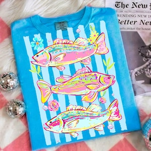 May include: A blue t-shirt with a vertical striped pattern and three colorful fish illustrations. The fish are pink, yellow, and green, with floral accents. The shirt has a Comfort Colors tag.