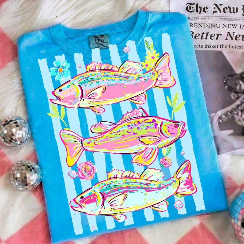 May include: A blue t-shirt with a vertical striped pattern and three colorful fish illustrations. The fish are pink, yellow, and green, with floral accents. The shirt has a Comfort Colors tag.