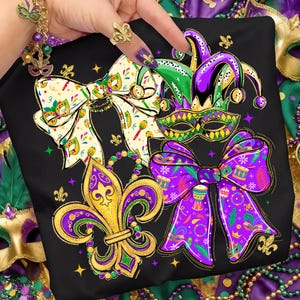 May include: Black t-shirt featuring a vibrant Mardi Gras design. The design includes a fleur-de-lis, bow ties, a jester hat, and a mask, all in purple, green, and gold. The shirt is decorated with stars and beads.