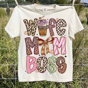 May include: Cream-colored t-shirt with the words "Wife Mom Boss" in various decorative fonts. The design includes a coffee drink illustration, leopard print, pink stripes, and a brown bow. The shirt is hanging on a clothesline.