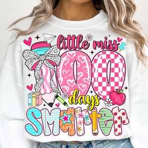 May include: White sweatshirt with a colorful graphic design. The design includes the text "Little Miss 100 Days Smarter" with hearts, flowers, and a pencil. The number "100" is in pink with heart and checkerboard patterns.