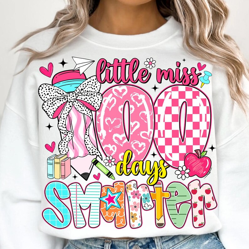 May include: White sweatshirt with a colorful graphic design. The design includes the text "Little Miss 100 Days Smarter" with hearts, flowers, and a pencil. The number "100" is in pink with heart and checkerboard patterns.