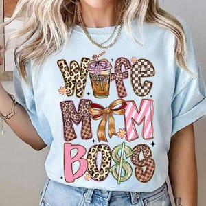 May include: Light blue t-shirt with the words "Wife Mom Boss" in a mix of leopard print, checkered, and pink lettering. A graphic of a drink with a straw is in the center. The shirt is a casual style.