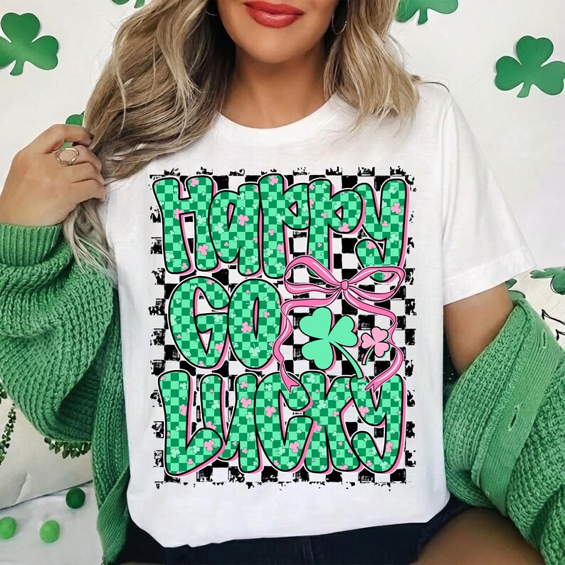 May include: White t-shirt with a St. Patrick's Day graphic. The design features the words "Happy Go Lucky" in green checkered letters, a pink bow, and a green shamrock. The shirt is a casual, festive apparel item.