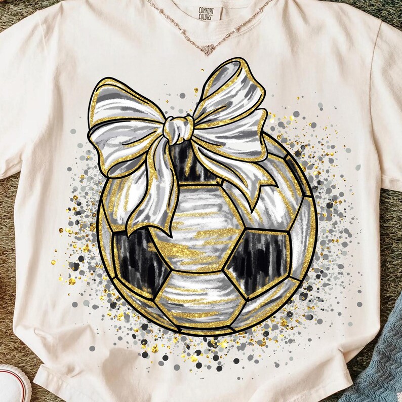 K&ouml;nnte beinhalten: Cremefarbenes T-Shirt mit einem Fu&szlig;ball-Design mit einer gro&szlig;en silbernen und wei&szlig;en Schleife. Der Fu&szlig;ball hat schwarze und goldene Akzente, mit goldenen Glitzerdetails und einem Spritzeffekt.