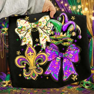 May include: Black tote bag featuring Mardi Gras themed designs. The bag displays a fleur-de-lis, bow ties, a jester hat, and a mask in gold, purple, green, and white. The bag is held by a person.