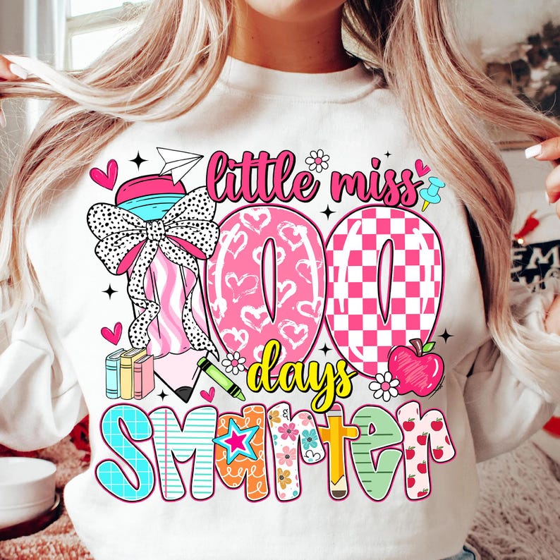 May include: White sweatshirt with a colorful graphic design. The design includes the text "little miss 100 days smarter" with pink hearts, a checkered pattern, and a cartoon apple. The sweatshirt has a crew neck.