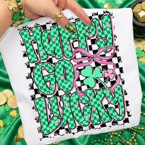 May include: White t-shirt with the text "Happy Go Lucky" in green checkered letters with pink shamrocks. A pink ribbon and green shamrock graphic are also featured on the shirt. The background has a black and white checkered pattern.