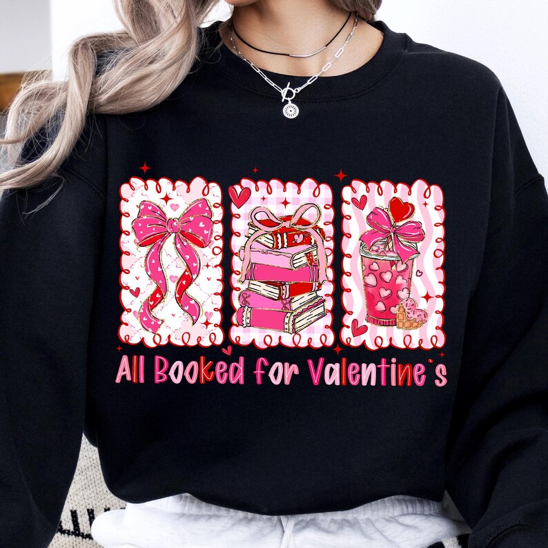 May include: Black sweatshirt with a Valentine's Day design. The design features three framed images: a pink bow, a stack of books, and a pink drink, all with hearts. The text "All Booked for Valentine's" is below.