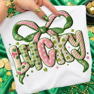 以下が含まれることがあります： 緑、ピンク、白で「LUCKY」の文字が刺繍された白いTシャツ。文字はクローバーとリボンで飾られています。上部には大きな緑とピンクのリボンがあります。シャツは、金貨とクローバーの装飾が施された緑色の背景にあります。