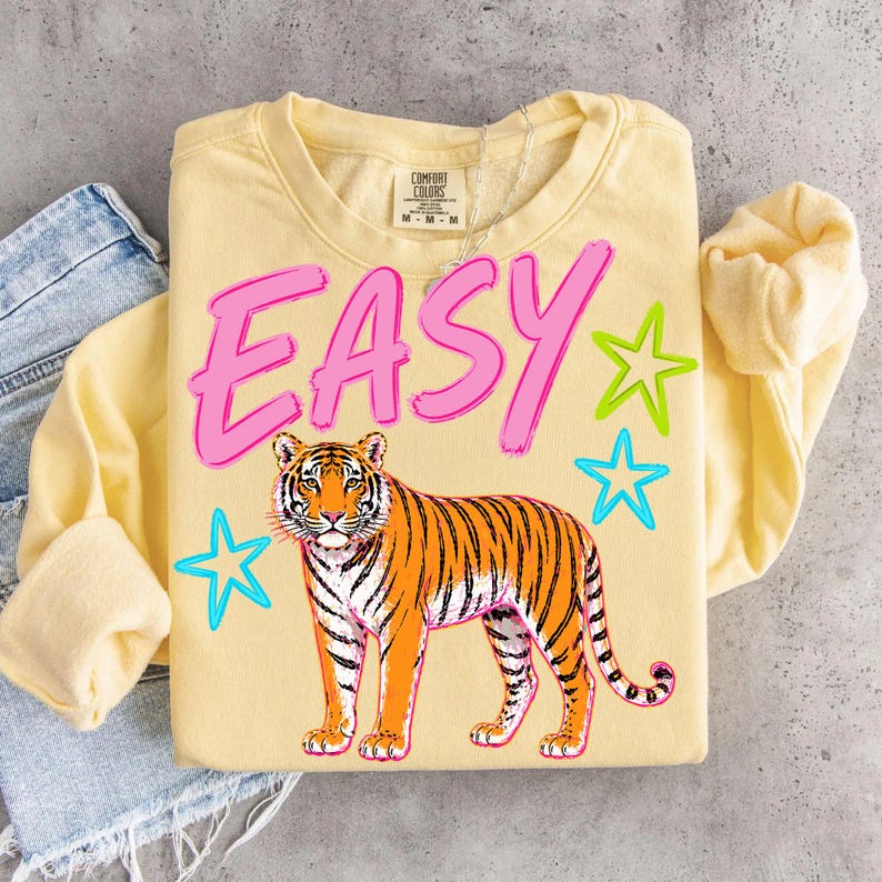 May include: A light yellow sweatshirt with the word "EASY" in pink block letters. Below the text is an orange and black tiger illustration, with green and blue star graphics.