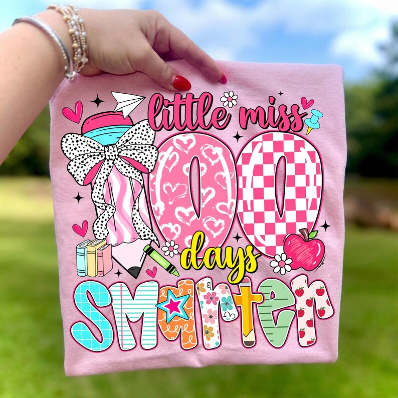 May include: Pink t-shirt with a colorful graphic design. The design includes the text "little miss 100 days smarter" with hearts, a bow, an apple, and other school-themed illustrations. The shirt is held up by a person.