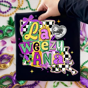 May include: Black t-shirt with the text "La Weezy Ana" in yellow, green, and purple block letters. The design includes a disco ball heart, a crown, and a fleur-de-lis, all in a Mardi Gras theme.