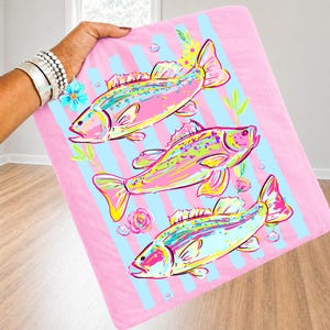 May include: A pink blanket with a repeating pattern of colorful fish, blue stripes, and floral accents. The fish are in shades of pink, yellow, and blue. The blanket is held up by a person.