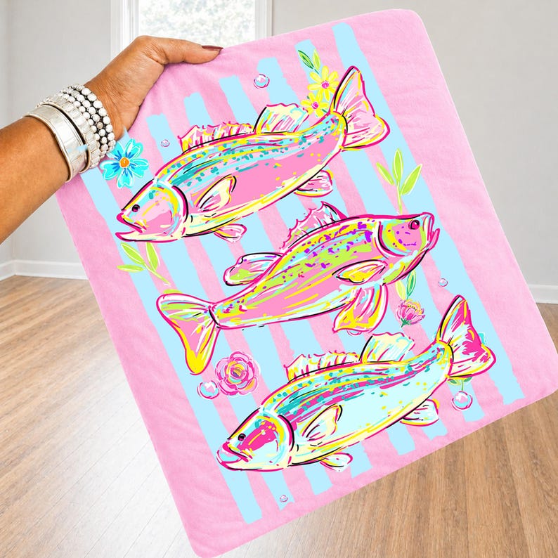 May include: A pink blanket with a repeating pattern of colorful fish, blue stripes, and floral accents. The fish are in shades of pink, yellow, and blue. The blanket is held up by a person.