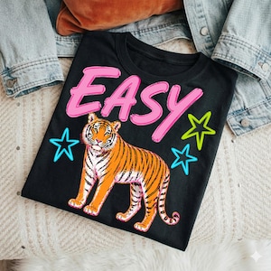 May include: Black t-shirt with a tiger graphic and the word "EASY" in pink. The tiger is orange and black with white stripes. Blue and green stars are also on the shirt.