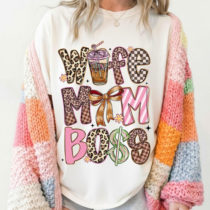 May include: Cream-colored graphic tee with the words "Wife Mom Boss" in various fonts and patterns, including leopard print, checkered, and pink stripes. A coffee cup graphic is also featured. The person is wearing a colorful, striped cardigan.