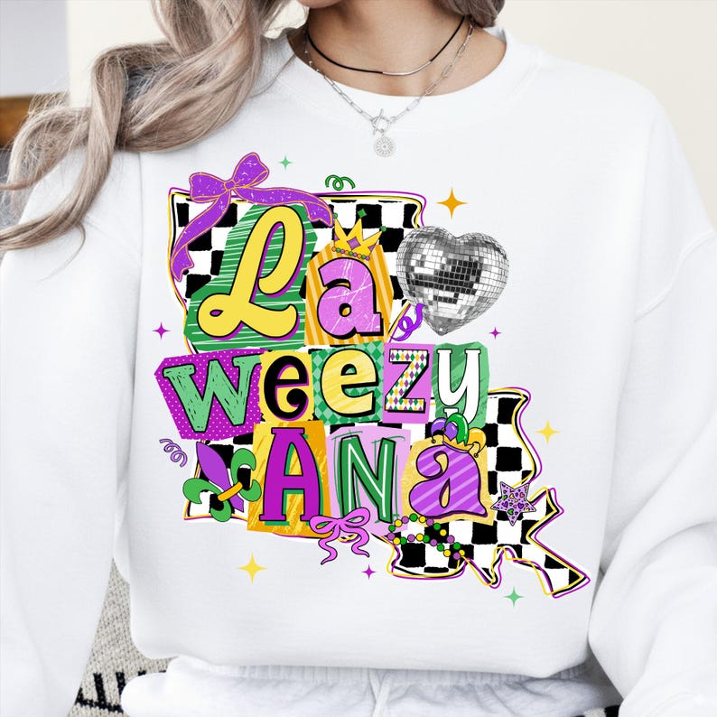 May include: White sweatshirt with a colorful graphic design. The design features the words "La Weezu Ana" in yellow, green, and purple, with a disco ball heart and a bow. The design is surrounded by stars and a checkered pattern.