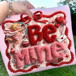 Peut inclure: Un coussin rose sur le th&egrave;me de la Saint-Valentin avec les mots "Be Mine" dans un design textur&eacute;. Il pr&eacute;sente un n&oelig;ud rouge, un cupidon avec un arc et des fl&egrave;ches, des c&oelig;urs, un cupcake et une lettre d'amour. La bordure est rouge et blanche.
