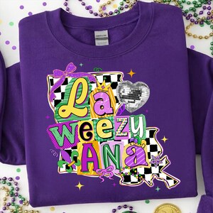 May include: A purple sweatshirt with a colorful graphic that says "La Weezu Ana". The design includes a disco ball heart, a crown, and a fleur-de-lis, with a black and white checkered pattern. The sweatshirt is decorated with Mardi Gras beads.