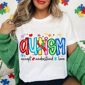 May include: White t-shirt with the word "AUTISM" in colorful, bubble letters. The shirt includes the words "accept, understand, love" below the main text, along with various graphics like hearts, flowers, and puzzle pieces.