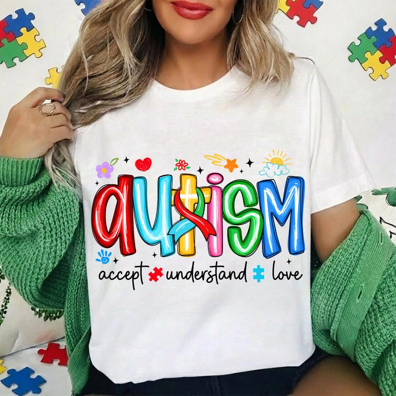 May include: White t-shirt with the word "AUTISM" in colorful, bubble letters. The shirt includes the words "accept, understand, love" below the main text, along with various graphics like hearts, flowers, and puzzle pieces.