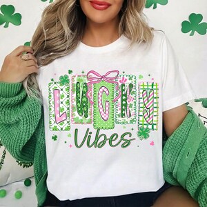 Op de afbeelding: Wit T-shirt met de woorden "LUCKY Vibes" in groen en roze, versierd met klavers en een roze strik. Het ontwerp heeft het thema St. Patrick's Day.