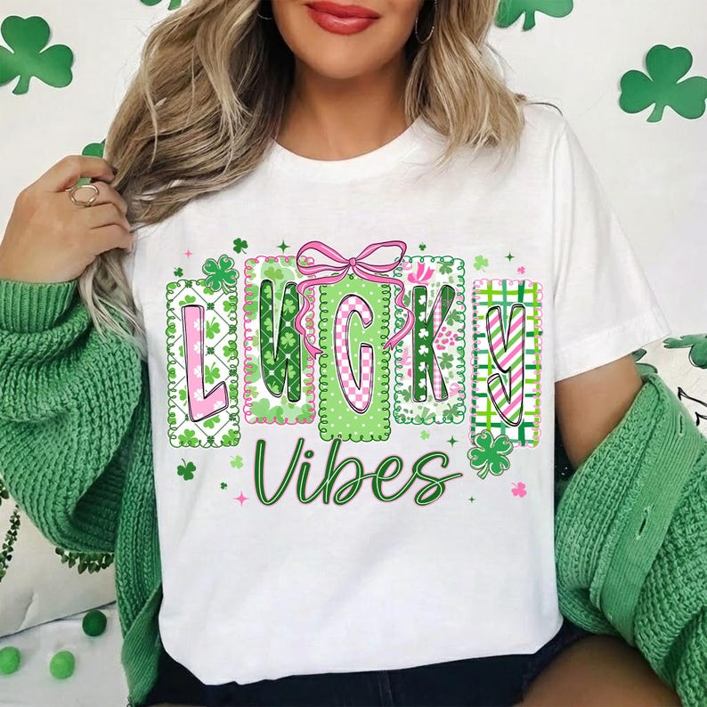 Op de afbeelding: Wit T-shirt met de woorden "LUCKY Vibes" in groen en roze, versierd met klavers en een roze strik. Het ontwerp heeft het thema St. Patrick's Day.