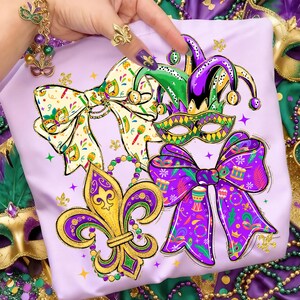 May include: A light purple t-shirt featuring Mardi Gras themed graphics. The design includes a fleur-de-lis, bow ties, a jester hat, and a mask. The graphics are in gold, green, and purple. The shirt is decorated with gold glitter.