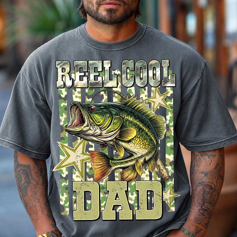 May include: Gray t-shirt with a graphic of a green and yellow fish with the text "REEL COOL DAD". The design includes camouflage and star accents, perfect for fishing enthusiasts.