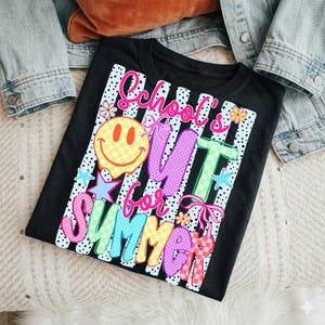 Puede incluir: Camiseta negra con la frase colorida "School's Out for Summer". El dise&ntilde;o incluye una carita sonriente, estrellas, flores y lazos. La camiseta se muestra sobre una superficie clara con una chaqueta vaquera.
