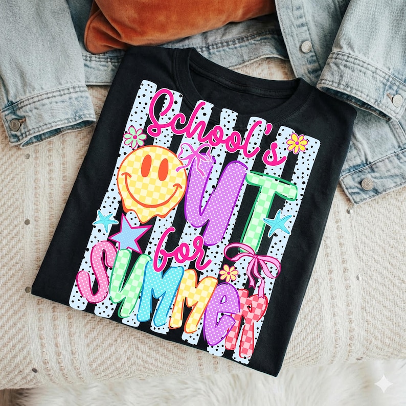 Puede incluir: Camiseta negra con la frase colorida "School's Out for Summer". El dise&ntilde;o incluye una carita sonriente, estrellas, flores y lazos. La camiseta se muestra sobre una superficie clara con una chaqueta vaquera.