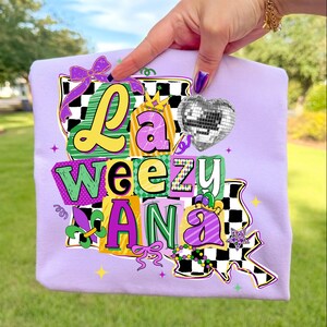 May include: A light purple t-shirt with the text "La Weezy Ana" in a colorful, blocky design. The design includes a disco ball heart, a crown, and a fleur-de-lis, with a checkered pattern and bow accents.