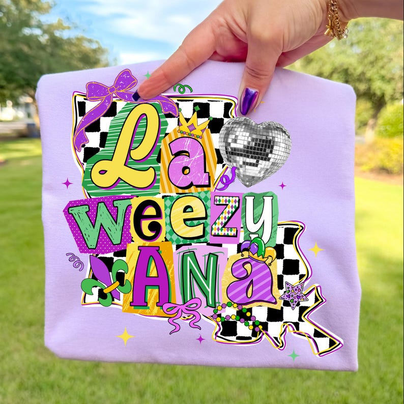May include: A light purple t-shirt with the text "La Weezy Ana" in a colorful, blocky design. The design includes a disco ball heart, a crown, and a fleur-de-lis, with a checkered pattern and bow accents.