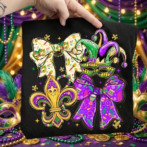 May include: Black tote bag featuring Mardi Gras themed designs. The bag displays a fleur-de-lis, bow ties, a jester hat, and a mask in gold, green, and purple. The bag is held up by a hand, with a background of Mardi Gras decorations.