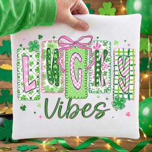 Op de afbeelding: Wit kussen met de woorden "LUCKY VIBES" in groen schrift. Het woord "LUCKY" staat in afzonderlijke blokken met roze en groene patronen. Boven de letter "G" staat een roze strik. Groene klavers en roze sterren zijn verspreid.
