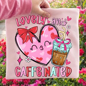 Puede incluir: Una sudadera rosa claro con un diseño gráfico. Presenta un corazón rosa con un lazo rojo, corazones y una cara sonriente. Una bebida de café está al lado del corazón. Las palabras "LOVELY and CAFFEINATED" están impresas en la sudadera.