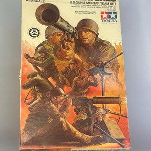 以下が含まれることがあります： タミヤ製の1/35スケールプラスチックモデルキット、アメリカ軍の迫撃砲と砲兵チームセットの箱。箱のイラストは、迫撃砲、機関銃、ライフルを持った戦闘中の兵士を描いています。箱は茶色で、黄色の文字とタミヤのロゴがあります。