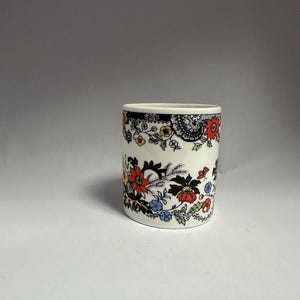 May include: A cylindrical ceramic cup with a floral pattern. The cup features a white base with a black rim and is adorned with red, blue, and yellow flowers and green leaves. The design wraps around the cup.
