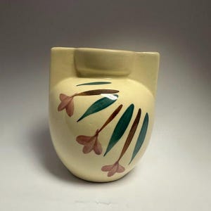 May include: Cream-colored ceramic vase with a rounded body and a rectangular top. The vase is decorated with pink flower and green leaf designs. The vase has a small, circular opening on the top.