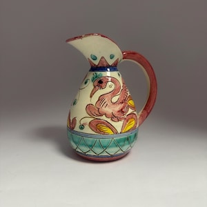 May include: A small, hand-painted ceramic jug with a cream body and a red handle. The jug features a pink bird design and is decorated with blue, yellow, and green accents. The base has a turquoise and red geometric pattern.