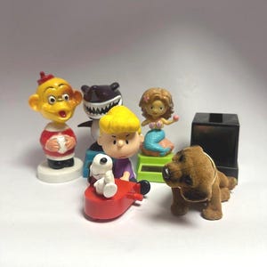 May include: A collection of small figurines, including a monkey, a shark, a mermaid, a dog, and Charlie Brown with Snoopy on a red plane. The figurines are brightly colored and made of plastic.
